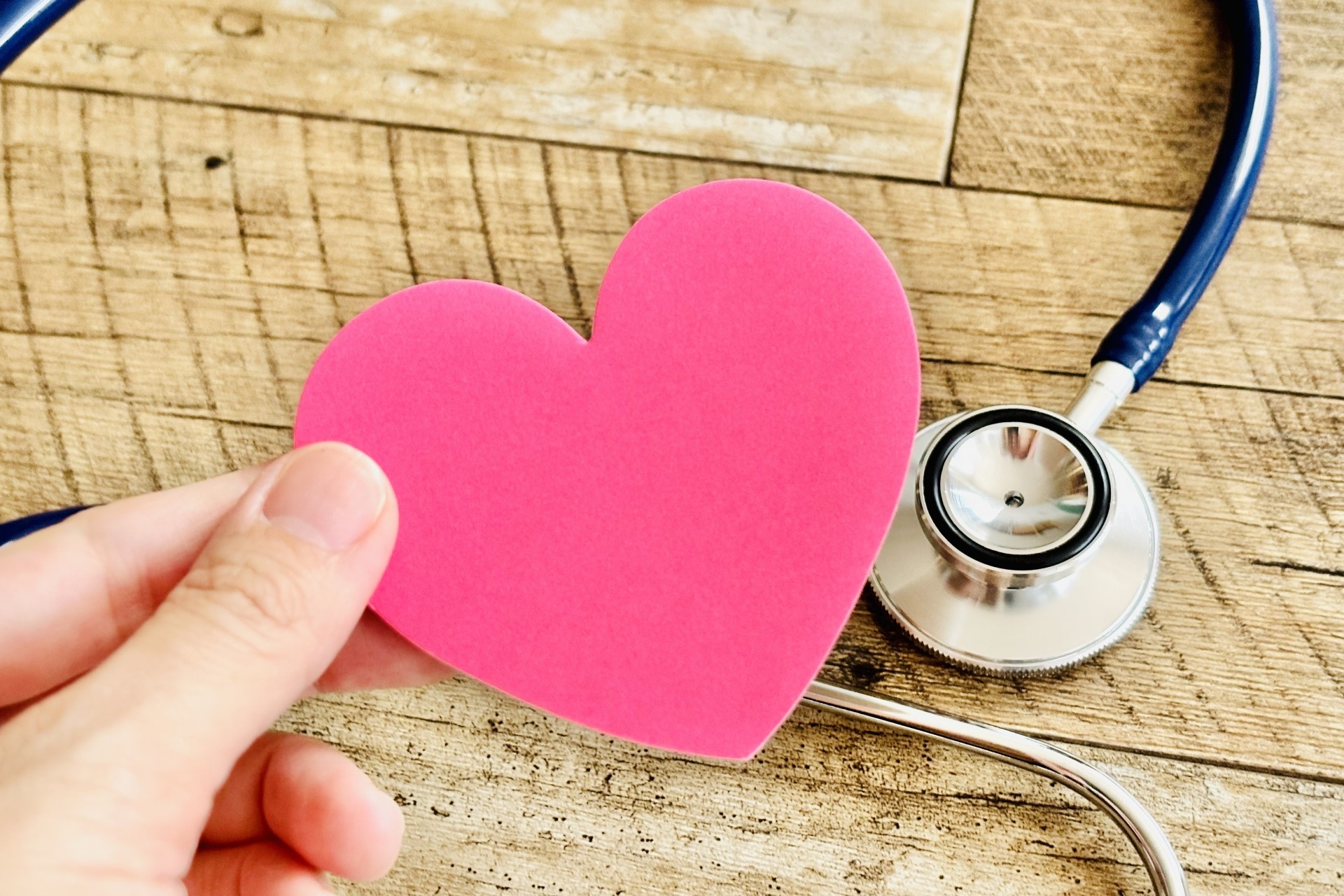 机の上に聴診器が置かれ、その上にピンクの紙のハートを持つ手がある