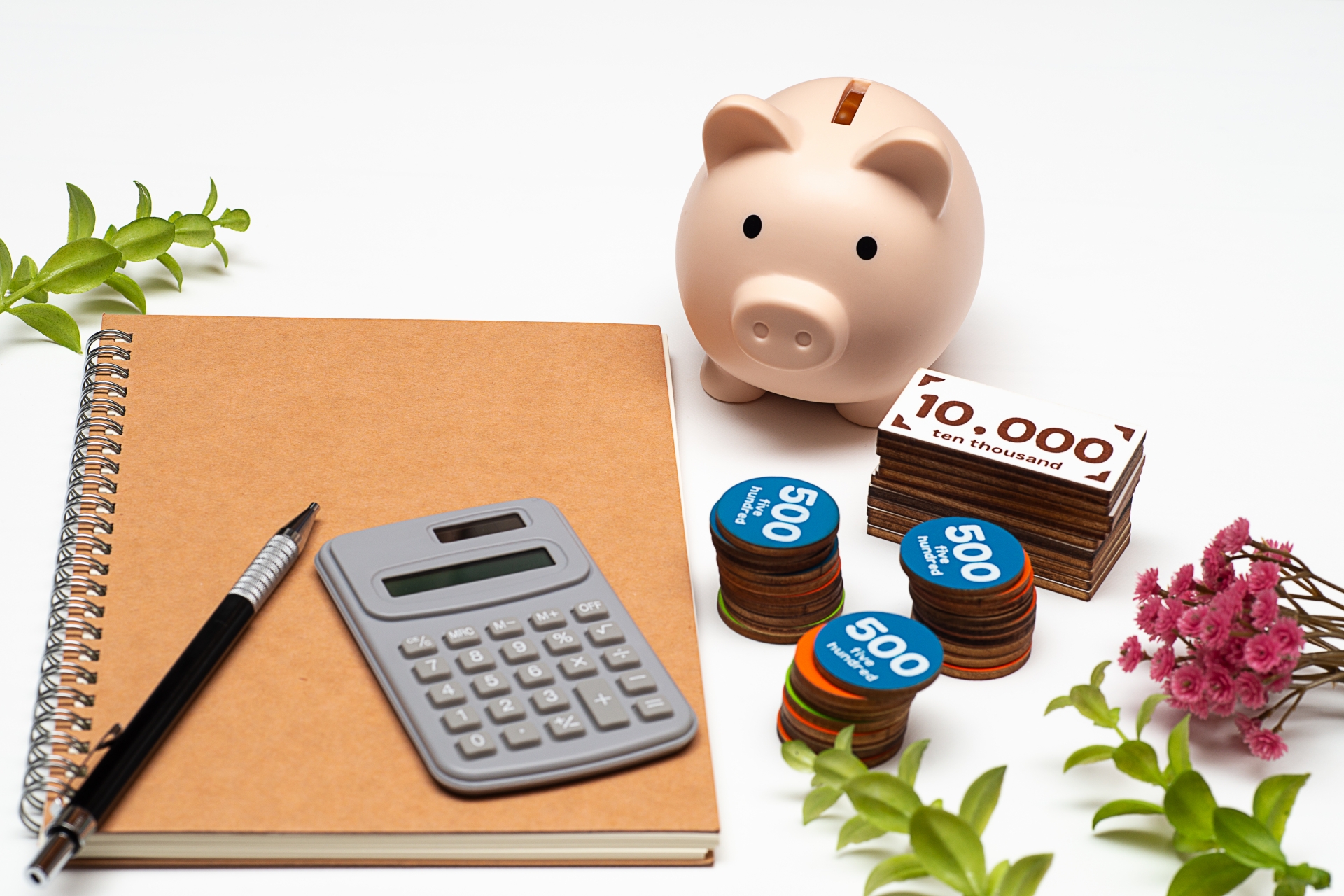 机の上にノート、電卓、ペン、おもちゃのお金、豚の貯金箱が置かれている様子