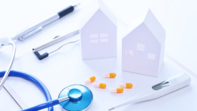 聴診器、ペン、カプセル、体温計、紙でできた家が机の上に乗っている