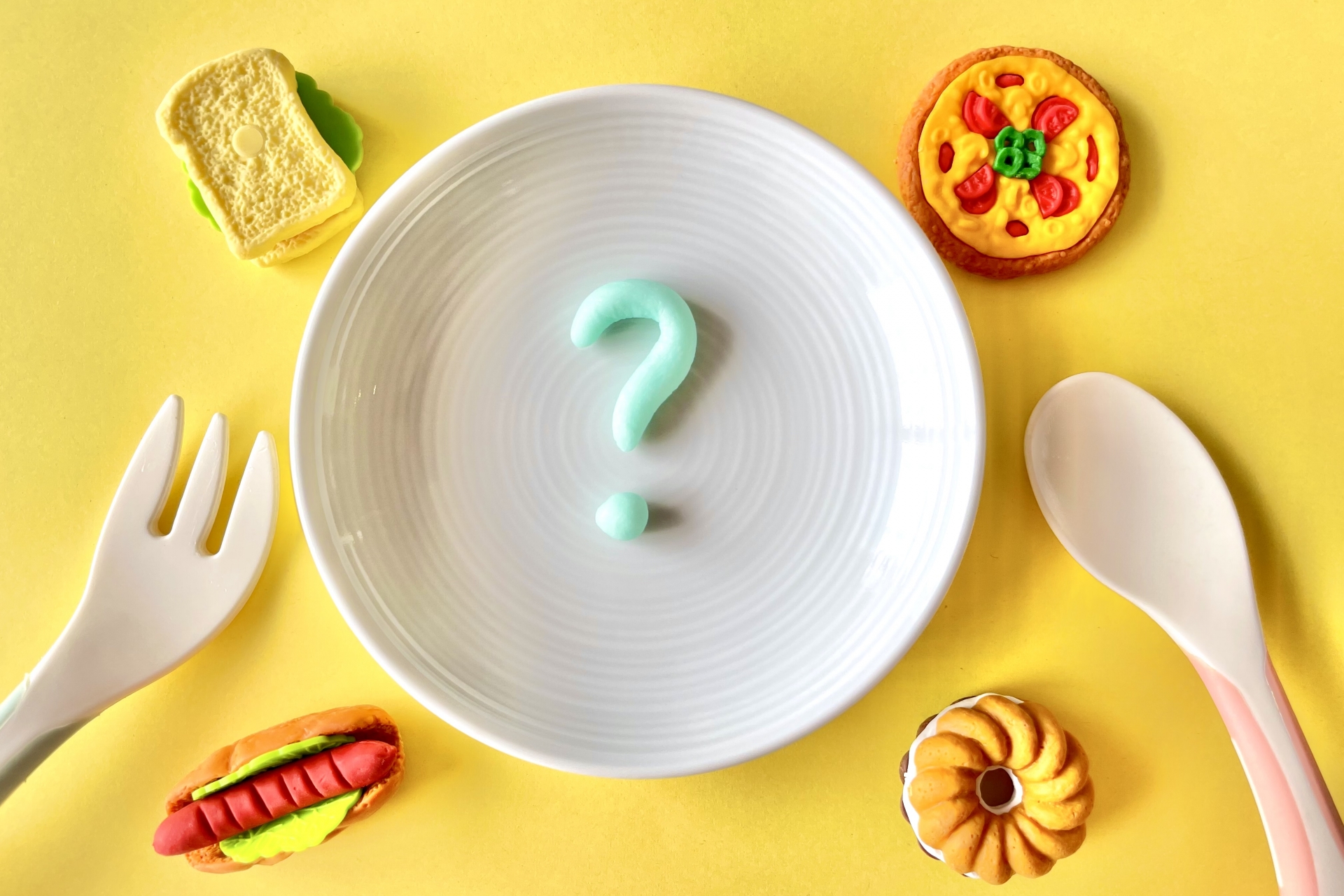 皿の上に粘土でできた?マークが置かれている様子。皿の周りにはサンドイッチ、ピザ、ドーナツ、ホットドッグのおもちゃが置かれている